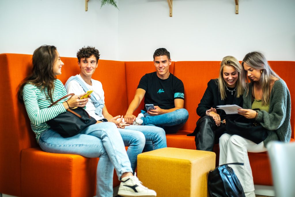 Een groep studenten zit samen op een oranje bank tijdens een meeloopdag op EuroCollege. Ze lachen en praten informeel met elkaar.
