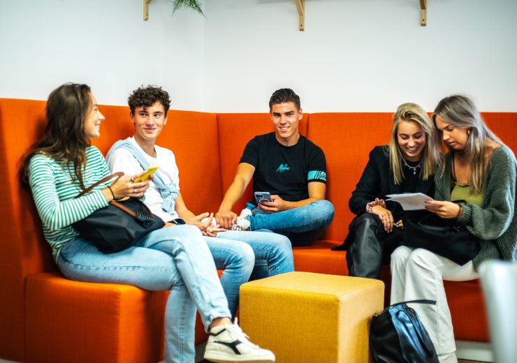 Een groep studenten zit samen op een oranje bank tijdens een meeloopdag op EuroCollege. Ze lachen en praten informeel met elkaar.